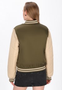 Giacca bomber verde oliva e beige con polsini e orlo a righe, caratterizzata da una texture liscia e una vestibilità affusolata.