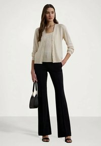 Mulher a usar um cardigan bege de malha canal e uma t-shirt de alças, calças largas pretas, segurando uma bolsa preta, em pé com uma mão no bolso.