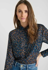 Long-sleeve, sheer blouse featuring a blue and brown animal print. Button-up design with a collar and two front pockets. Denim background.