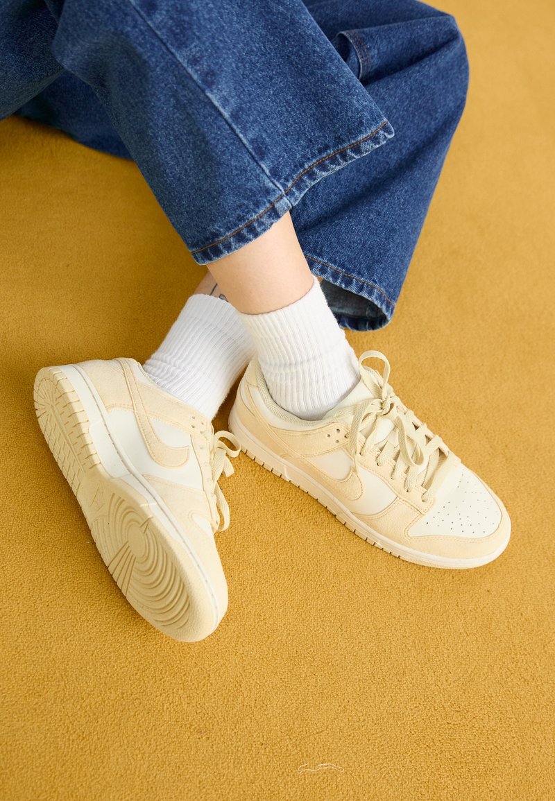 Beige Wildleder- und Leder-Sneaker mit einer strukturierten Gummisohle, kombiniert mit weißen gerippten Socken, die auf einer gelben Oberfläche ruhen. Jeans sind sichtbar.