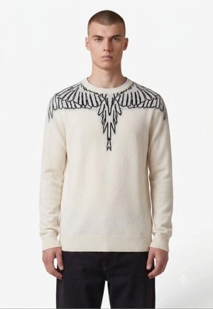 Giovane uomo che indossa un maglione color crema con un disegno di ali nere sulle spalle e sul petto, abbinato a pantaloni neri.