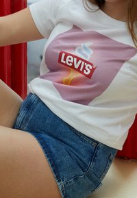 Osoba v bílém tričku Levi's s růžovým a žlutým designem a modrých džínových šortkách, sedící na pozadí v červené barvě.