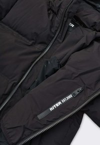 Giacca imbottita nera con chiusura a zip. Presenta una targhetta con logo, tasche con zip e tessuto leggero. Texture liscia e design minimalista.