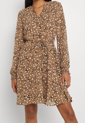 Femme portant une robe beige avec des taches abstraites noires et blanches, manches longues avec poignets élastiques, un col en V et une ceinture nouée à la taille.