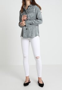 Hellgraues Langarm-Hemd mit Knopfleiste und strukturiertem Muster, kombiniert mit zerrissenen weißen Skinny-Jeans und schwarzen spitz zulaufenden Ballerinas.