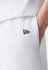 Tejido de sudadera gris claro con una textura suave, que presenta un pequeño logo negro bordado en el lado izquierdo. Diseño minimalista, corte relajado.