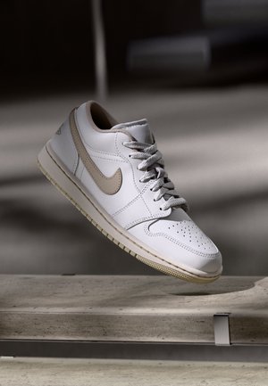 Biały i beżowy sneaker Nike unoszący się nad betonowym krawędzią z delikatnym cieniem w tle.