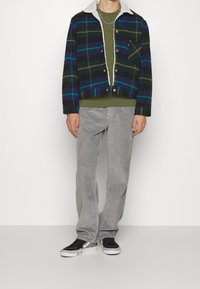 Chaqueta de franela a cuadros en azul marino, verde y amarillo con un cuello de borreguito blanco, combinada con un suéter verde y pantalones de pana grises. Zapatillas negras.