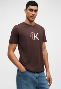 Hnedé bavlnené tričko s krátkymi rukávmi, na ktorom je biele a ružové logo Calvin Klein. Nosí sa s svetlo modrými džínsami, ruky v vreckách.