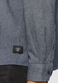 Blauwe spijkerblouse van een textuurstof, met lange mouwen en knoopmanchetten. Heeft een genaaide label aan de onderkant met de merknaam.