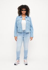 Chaqueta de mezclilla azul claro con dos bolsillos frontales. Lleva puesta sobre una blusa blanca y combinada con unos jeans azul claro. Zapatillas deportivas grises y naranjas.