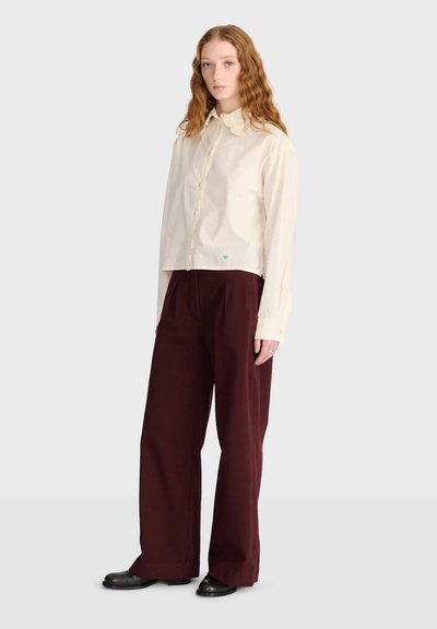 Femme aux longs cheveux ondulés portant un chemisier crème boutonné et un pantalon ample rouge foncé, debout devant un fond clair uni.