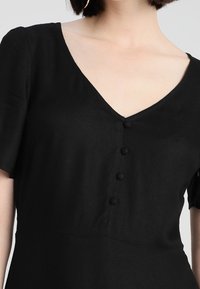 Robe noire avec un décolleté en V, manches courtes et cinq boutons sur le devant. Fabriquée en tissu doux avec une coupe décontractée.