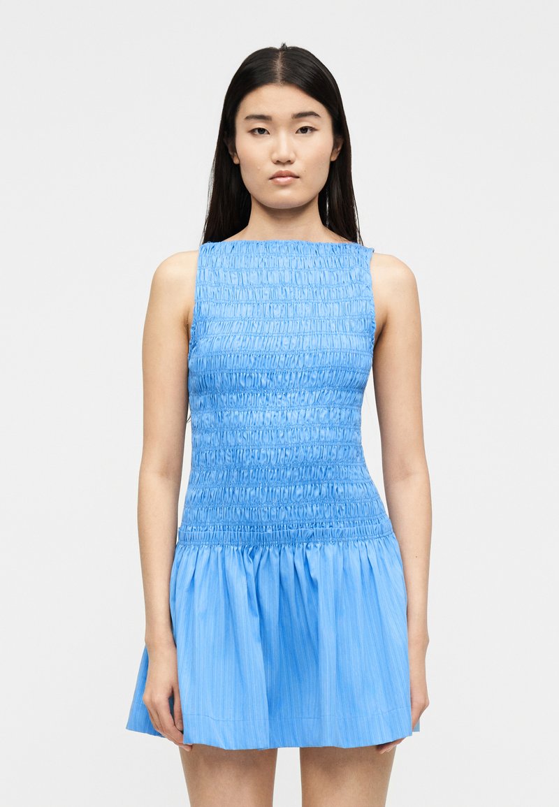 Νέα γυναίκα με μακριά σκούρα μαλλιά, φορώντας αμ άνικο, γαλάζιο μίνι φόρεμα με ρυτίδες, στέκεται μπροστά σε ένα απλό λευκό φόντο.