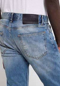 Pantalones vaqueros de mezclilla azul claro con una textura descolorida, con una parche de cuero negro en la cintura y dos bolsillos traseros con detalles de costura.