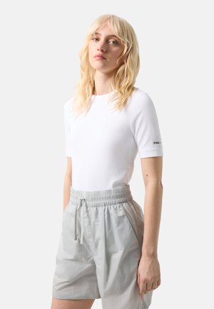 Blondynka ubrana w białą, prążkowaną bluzkę z krótkim rękawem i jasnoszare szorty z elastycznym pasem, stojąca na jednolitym tle.