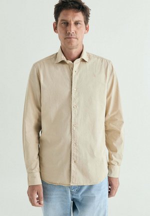 Hombre con camisa beige de manga larga abotonada y vaqueros azul claro, de pie frente a un fondo blanco liso.