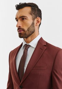 Brąsowy garnitur o eleganckim kroju, z klapami typu notch, pojedynczym zapięciem na guzik i pasującą chusteczką w kieszeni. Biała koszula i wzorzysta krawat.