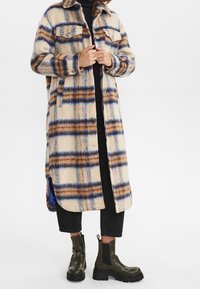 Person in einem langen beigen, blauen und braun karierten Mantel, schwarzen cropped Pants, schwarzem Oberteil und dunkelgrünen klobigen Stiefeln vor einem weißen Hintergrund.