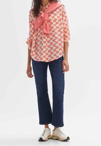 Long-sleeve shirt in coral wave pattern, paired with dark blue cropped jeans and white sneakers with colorful accents. Sweater tied around the neck.