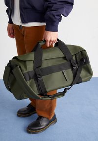 Olivengrønn duffelbag laget av vanntett materiale, med svarte stropper, et håndtak på toppen, og et minimalistisk design med logodetaljer.