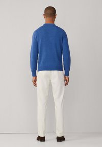 Blå stickad tröja med långa ärmar, rund hals och ribbade manschetter, ihop med vita byxor och bruna skor. Minimalistisk bakgrund.