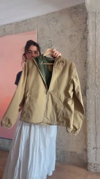 Femme tenant une veste beige zippée sur un cintre en bois devant un mur à texture rugueuse et un panneau rose.