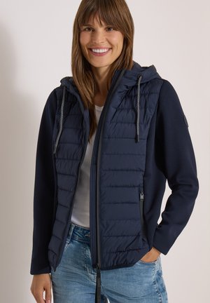 Smilende kvinde med brunt hår iført en navyquiltet hættejakke over en hvid skjorte og blå jeans, stående med den ene hånd i lommen.