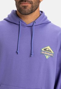 Fialová fleecová mikina s klokánkovou kapsou, kapucňou na šnúrku a farebnou nášivkou loga Quiksilver na hrudi. Mäkká textúra, voľný strih.
