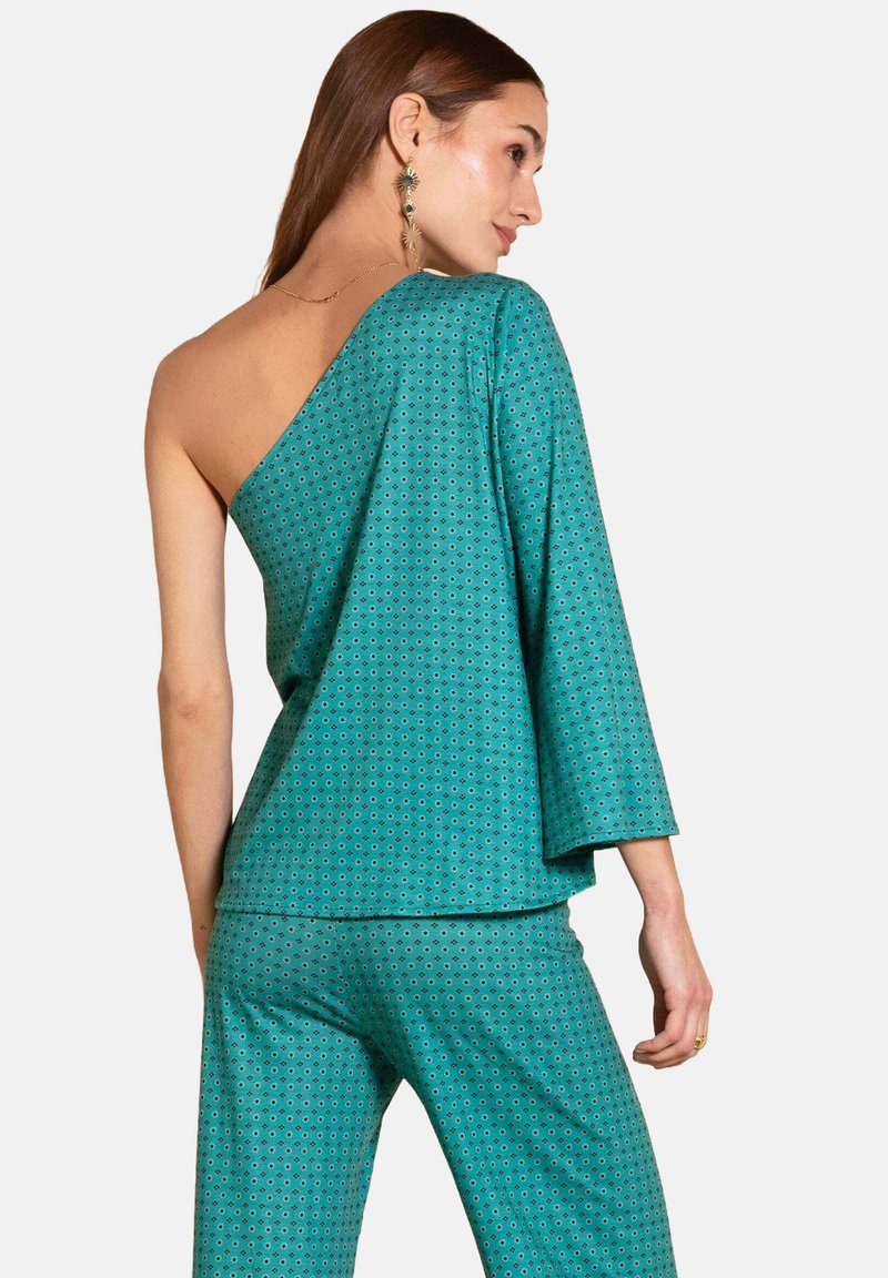 Femme portant un top asymétrique teal à motifs avec une large manche et un pantalon assorti, vue de dos contre un fond uni.