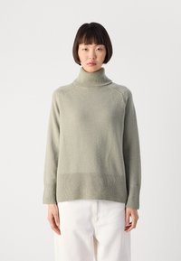 Woman with short black hair wearing a light gray turtleneck sweater and white pants, standing against a plain light background.