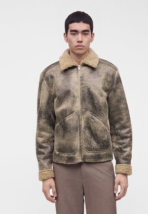 Young man wearing a faded brown shearling jacket with a fluffy collar and cuffs, standing against a plain light gray background.