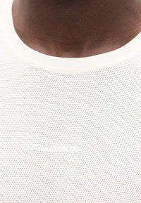 Κοντινό πλάνο ενός ατόμου που φοράει λευκό αθλητικό μπλουζάκι με οπές, στρογγυλή λαιμόκοψη και αχνό λογότυπο στο στήθος.