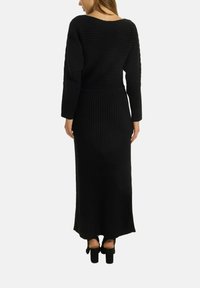 Schwarzes geripptes Strickkleid mit langen Ärmeln, weitem Ausschnitt und taillierter Taille, die zu einem fließenden Rock übergeht. Sichtbare klobige Absatzschuhe.