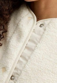 Close-up of a person wearing a cream-colored sherpa jacket with metal snap buttons and curly dark hair visible on the left side.