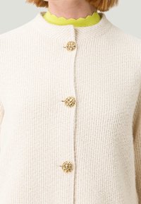 Cardigan in maglia color crema con collo rotondo e chiusura frontale con un unico bottone intrecciato dorato, con un vivace colletto giallo smerlato sotto.