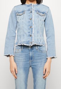 Mujer con chaqueta de mezclilla azul claro con bordes deshilachados y detalles de strass en los bolsillos, combinada con pantalones de mezclilla azul claro a juego.