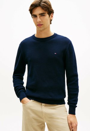 Junger Mann mit braunen Haaren, der einen marineblauen, strukturierten Pullover und beige Hosen trägt, steht mit einer Hand in der Tasche vor einem weißen Hintergrund.