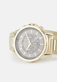 Reloj de acero inoxidable dorado con una esfera gris oscura, subesferas de cronógrafo, visualización de fecha y caja octagonal. Presenta marcadores de horas audaces y una correa texturizada.