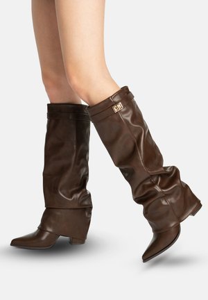 Jambes portant des bottes en cuir marron foncé, hautes jusqu'aux genoux, souples, avec des talons bas et un détail de boucle dorée, sur fond blanc.