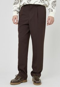 Pantalons plissés marron en tissu léger, présentant un design à jambe droite et des poches latérales, associés à des chaussures en cuir marron épais.