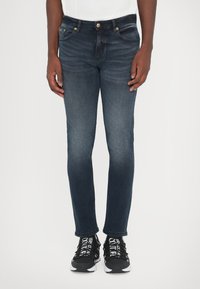 Σκούρο μπλε τζιν παντελόνι με στενή εφαρμογή, διαθέτοντας κλασικό σχέδιο πέντε τσεπών, εμφανή ραφή και χρυσά κουμπιά.