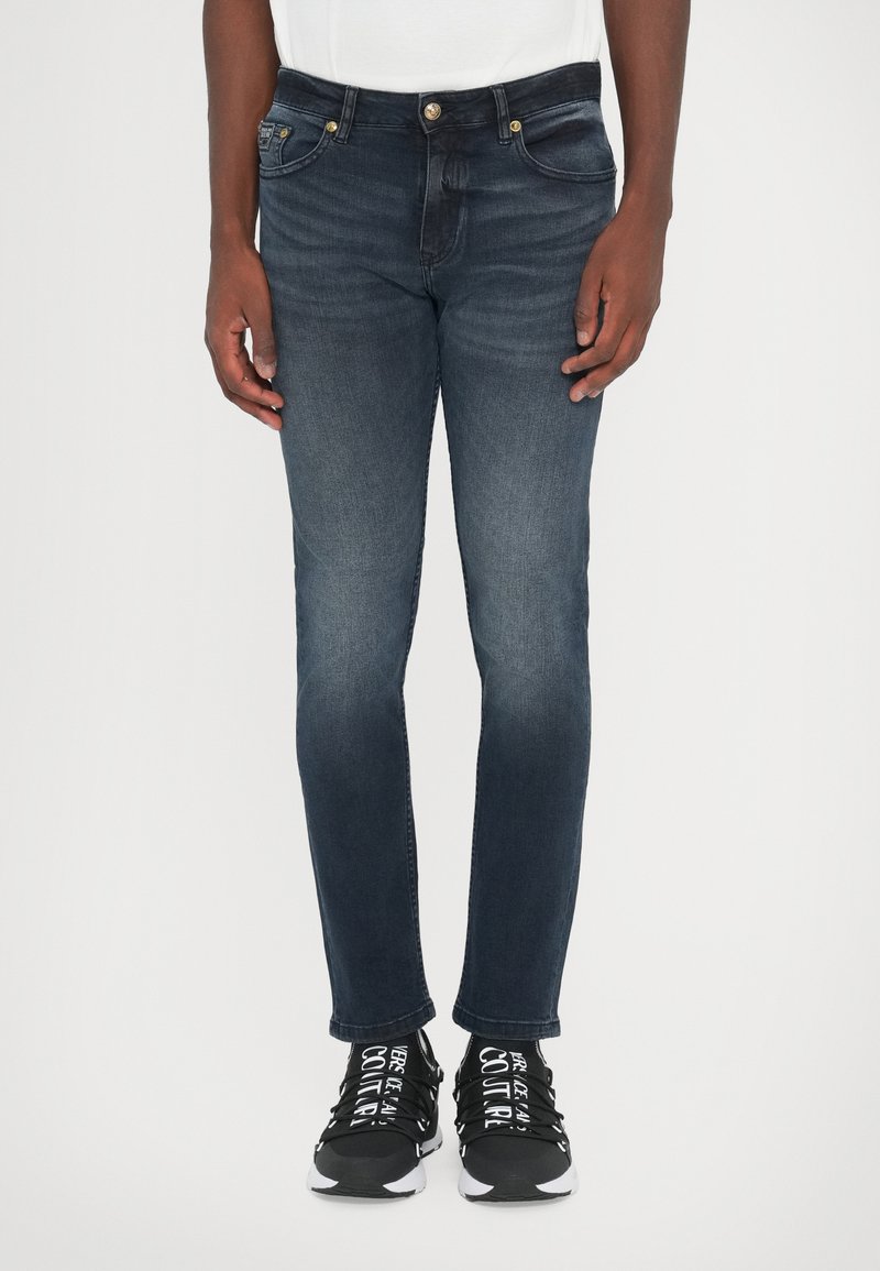 Σκούρο μπλε τζιν παντελόνι με στενή εφαρμογή, διαθέτοντας κλασικό σχέδιο πέντε τσεπών, εμφανή ραφή και χρυσά κουμπιά.