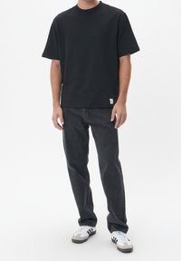 Camiseta negra de algodón con cuello redondo, combinada con jeans gris oscuro y zapatillas blancas con detalles en gris. La camiseta tiene una pequeña etiqueta en el dobladillo.