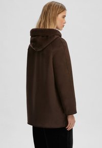 Cappotto marrone con cappuccio, dotato di un colletto morbido e testurizzato e linee pulite. Presenta una vestibilità rilassata e dettagli sulle cuciture laterali, realizzato in un materiale liscio.