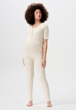 Schwangere Frau steht barfuß mit einem beigen, taillierten Oberteil mit kurzen Ärmeln und passenden Leggings vor einem schlichten, hellen Hintergrund.