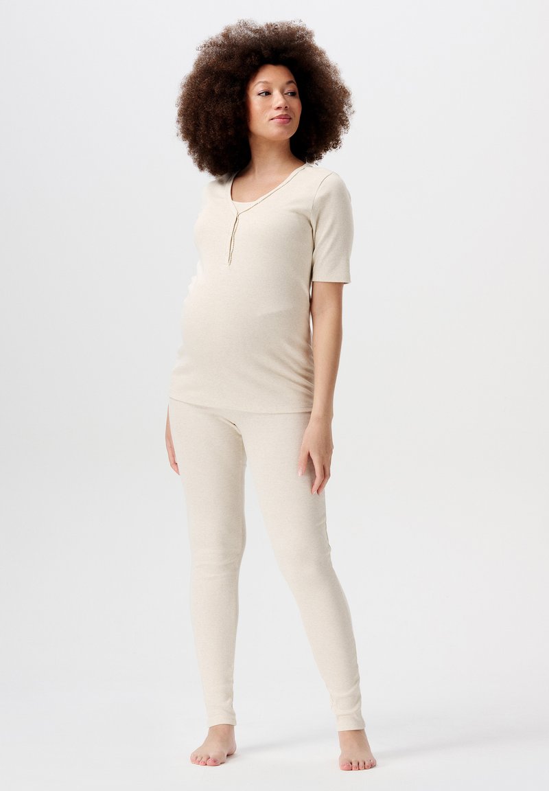 Gravid kvinna står barfota, iklädd en beige figurnära topp med korta ärmar och matchande leggings, framför en enkel ljus bakgrund.