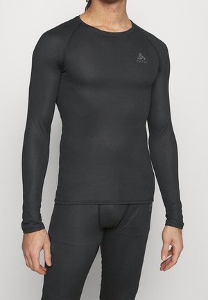 Mężczyzna ubrany w dopasowaną czarną termiczną bluzę z długim rękawem oraz dopasowane termiczne legginsy, stojący na tle jednolitego jasnego tła.