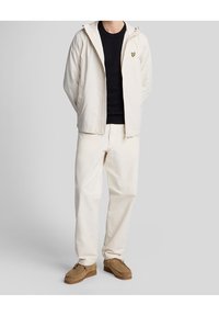 Helle beige Kapuzenjacke mit Reißverschluss und besticktem Logo, kombiniert mit cremefarbenen Hosen und braunen Wildlederschuhen, die ein lässiges Outfit ergeben.