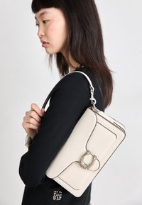 Sac à main beige clair au design structuré, doté d'un accent décoratif ovale en métal et d'une surpiqûre foncée contrastante. Bandoulière réglable.